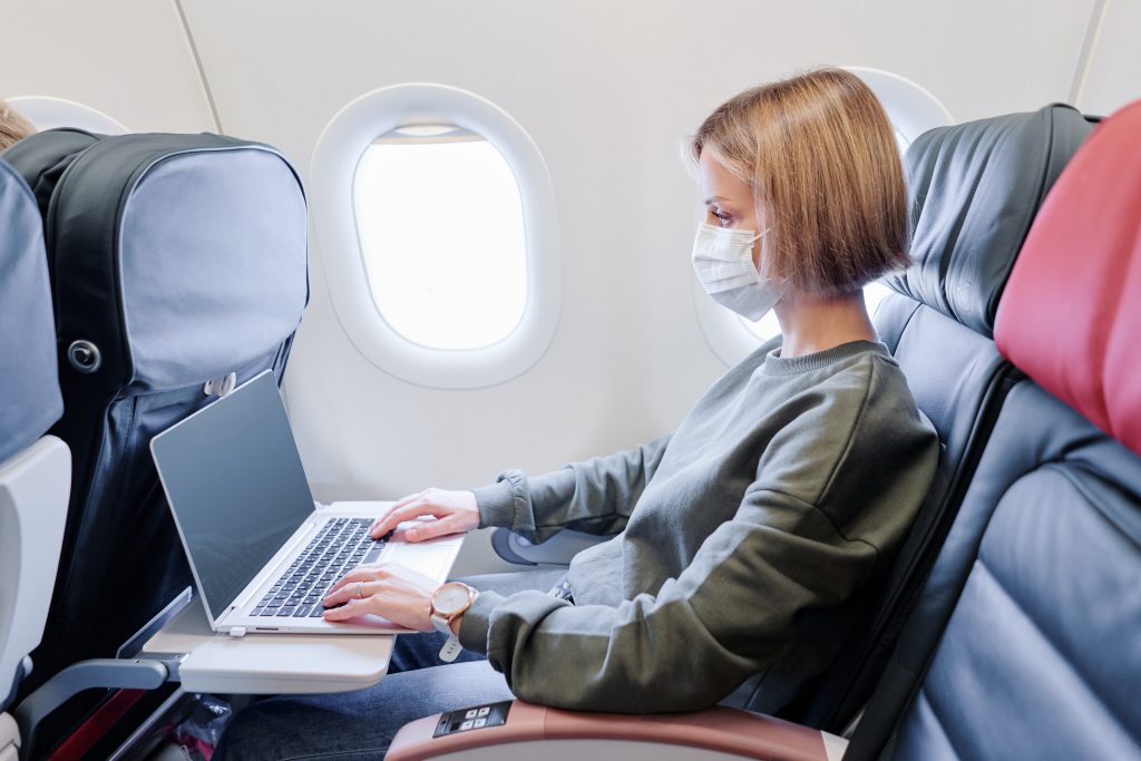 white blonde female in grey sweats wearing face mask sits in airplane seat using computer. One of the 7 tips to survive long international flights.