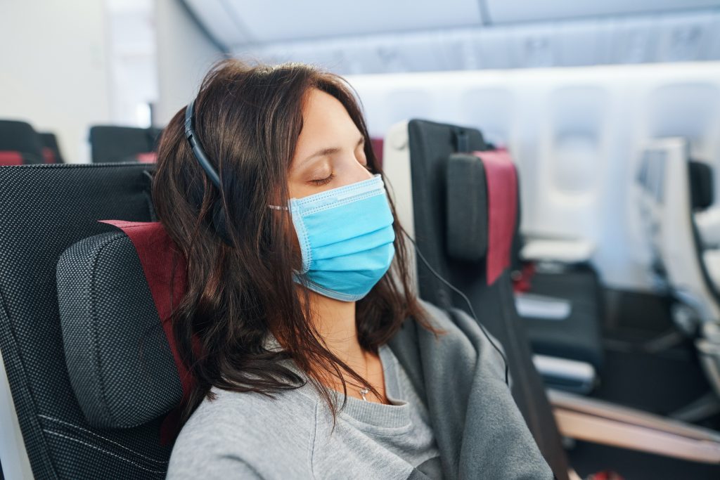 White female with dark hair wearing facemask sleeping on airplane.