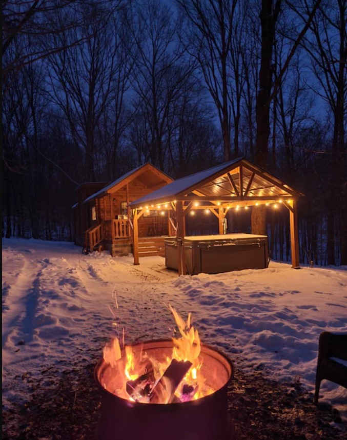 A log cabin, a gazebo with string lights, and a hot tub, and a fire pit in the woods at twilight. There is snow on the ground, the string lights are on, and there is a fire in the fire pit. 