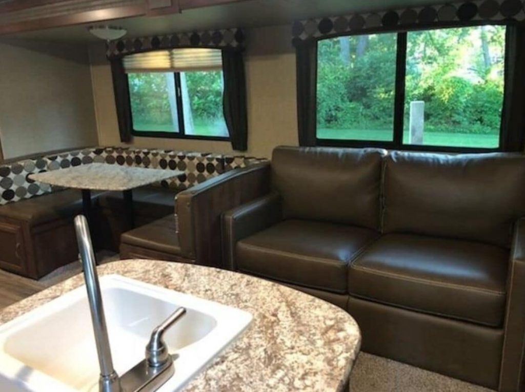 The living area of a luxury RV with a loveseat, a dining booth, kitchen, and lots of windows. 