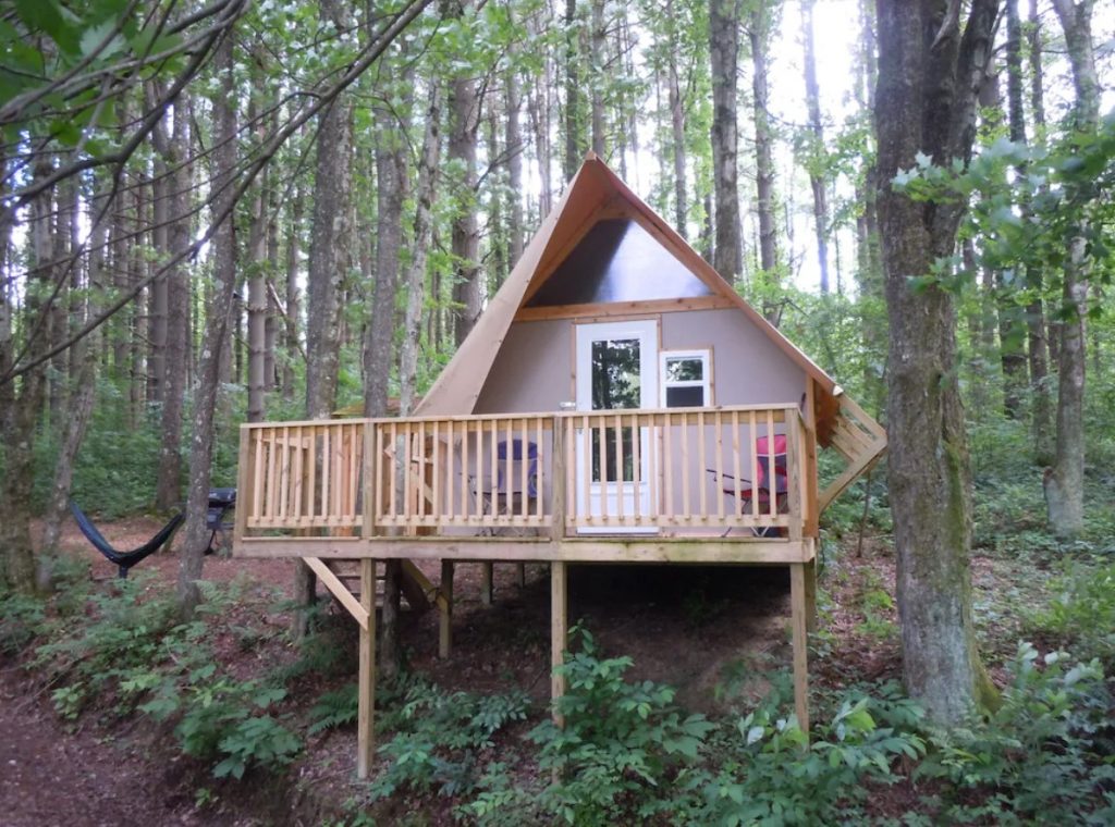 The front of a pine and canvas glamping cabin perched on the side of a wooded hill. It has a front porch. 