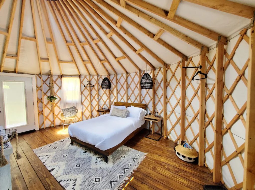 The inside of a large yurt where there is a bed with white sheets, black wicker lights, a patterned rug, and a large glass door. It's one of the best places for glamping in Ohio. 