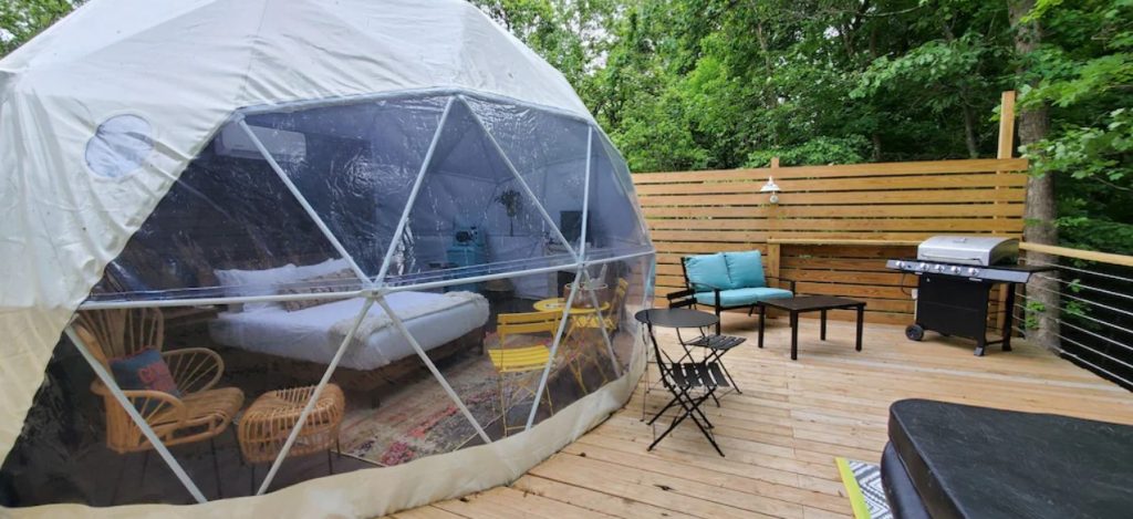 The exterior of a geodesic dome on a wooden platform. The front of the dome is clear so you can see furniture inside. On the platform there is seating and a grill. Its one of the best places for glamping in Ohio. 