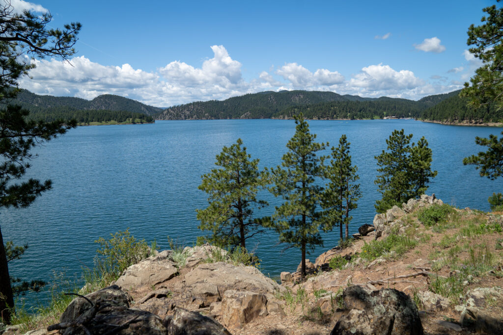 Large lake with bright blue water surrounded by tree-covered mountains with rocks and evergreen trees in the foreground. Things to do in Rapid City.