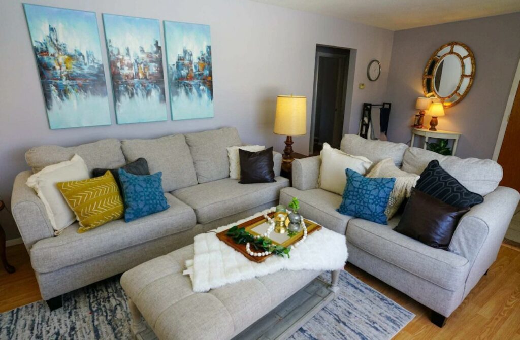 A cozy living room with two couches, an ottoman, and paintings on a light grey wall. 
