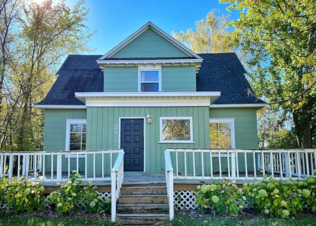 The exterior of a historic home that looks like a cottage. It is painted a blue-green color and there is a wrap around porch surrounded by trees and bushes. One of the best Airbnbs in North Dakota. 