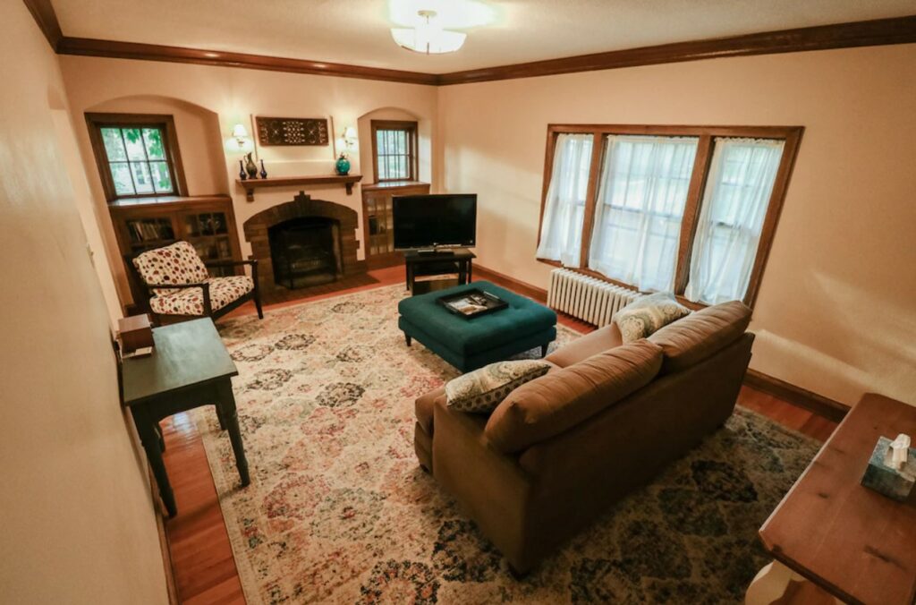 The cozy living room of a historic duplex that has a couch, an arm chair, a brick fireplace, and lots of windows. 