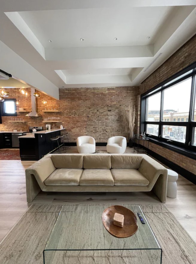 A modern studio apartment with exposed brick walls, a modern sofa and arm chairs, a modern kitchen, and large windows that look out onto a city