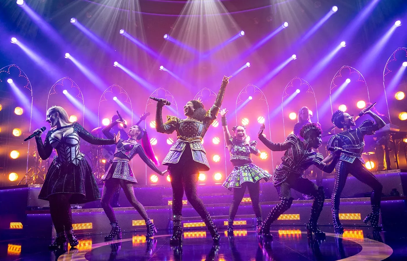 Six The Musical moment with 6 women singing with microphones in hand and lit with purple lighting.