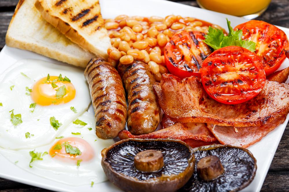 Close up a full English Breakfast on a plate with tomatoes, mushrooms, bacon, sausage, egg and beans.  