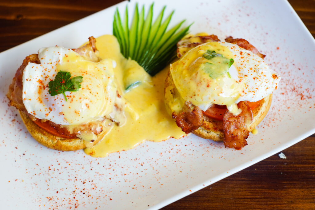 Eggs Benedict on a rectangle white plate. There is bacon and tomatoe under the egg and papriks sprinked on top. 