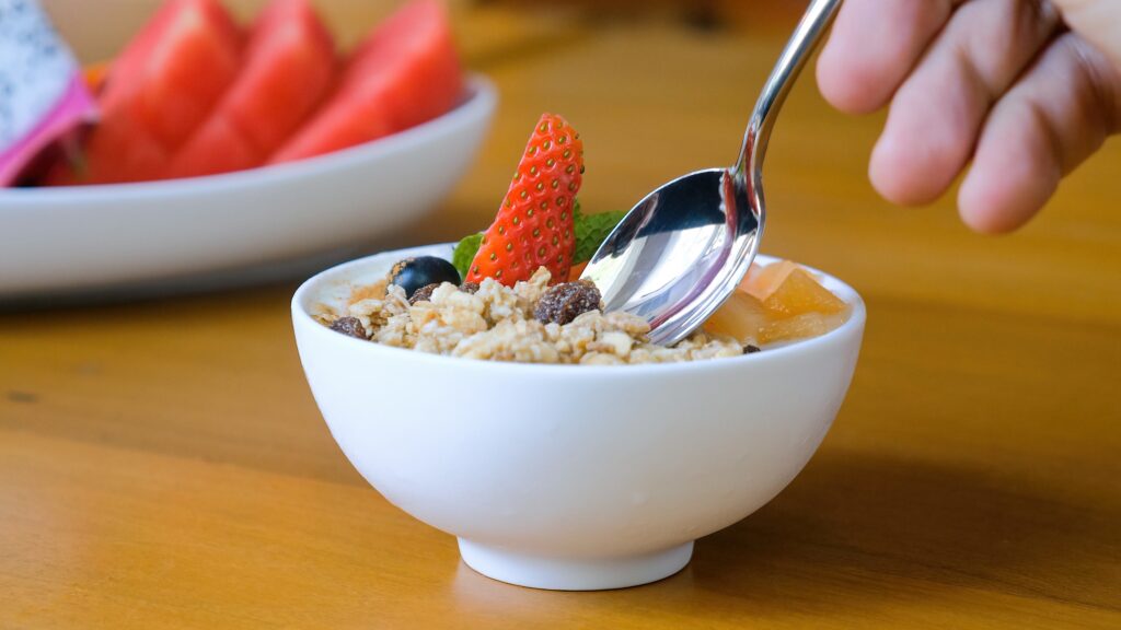 Human hand with spoon eat healthy breakfast food.  Bowl of homemade granola with yogurt, raisins, fresh berries, strawberry, melon, papaya on wooden table in restaurant