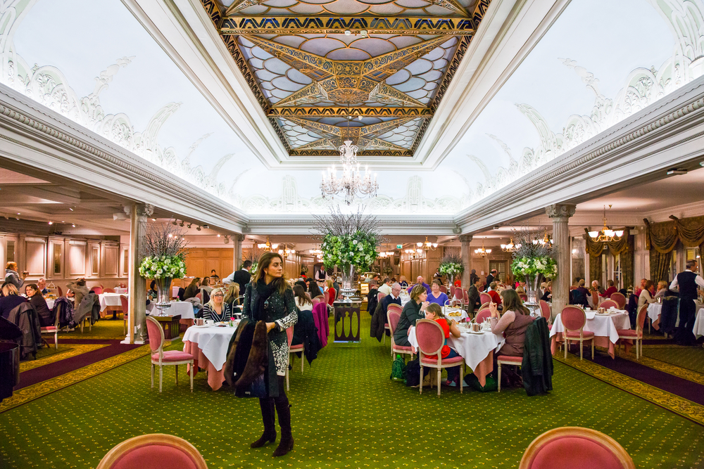 The dining room at Harrods with people sat at tables with green carpet and very ornate ceiling.