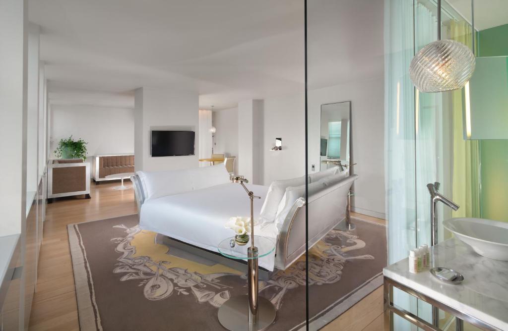 A beautiful white bedroom at the Sanderson Hotel. there is a glass wall, a white bed and a brown sofa in the distance. 