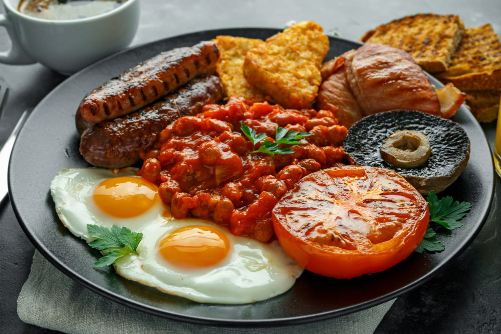 A plate of a full Irish breakfast with sausage, beans, eggs, white pudding, hashbrowns, a mushroom, roasted tomatoes, and toast.