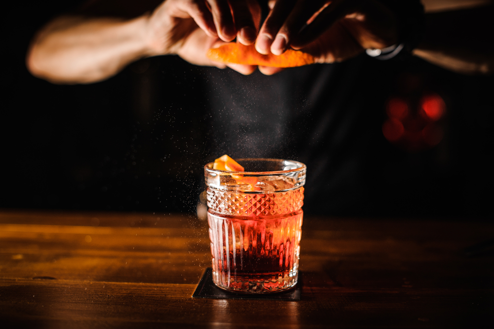 bartener making a cocktail. They are squeezing orange into a glass full of a cocktails. 