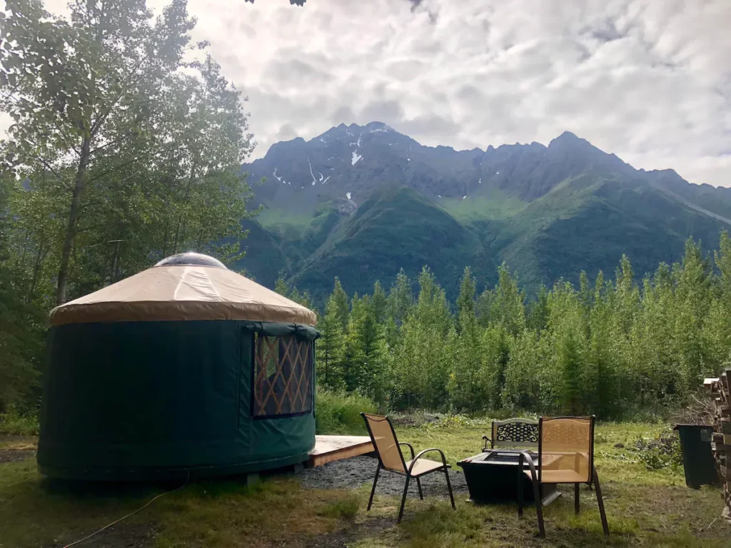 A Riverfront Yurt on the sides of a wooded area in front of mountins. The article is about camping in Alaska. 