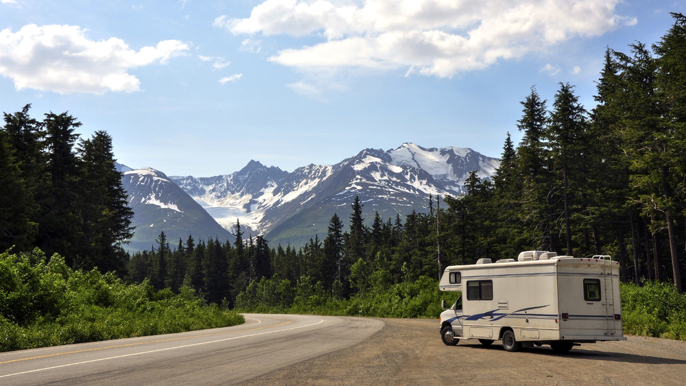 Motorhome by a road with mountains in the background.  This article is about camping in Alaska. 