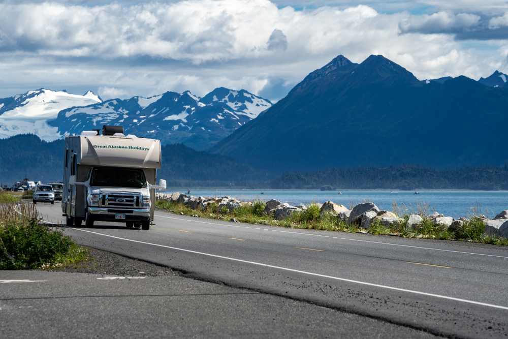 Recreational vehicle drives down the Homer Spit. There are mountains in the background and a lake to the side of the vechicle. 