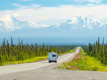 RV driving down road with mountains in background while camping in Alaska
