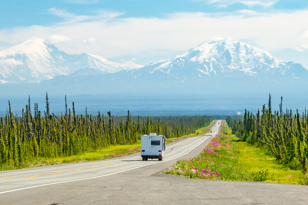 RV headed to the Alaska Range mountains on horizon at the end of long highway with green trees on side of highway. 