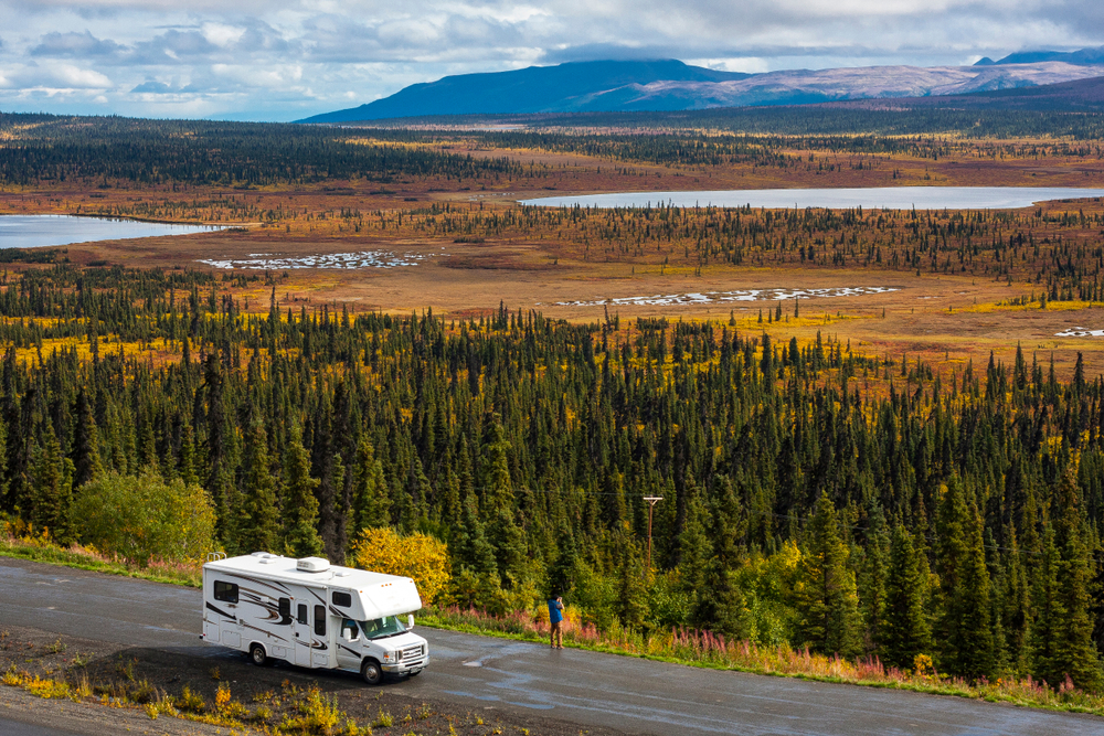 RV next to a road in a parking lot in Alaska with spectacular, beautfiul background with lakes and conifer forest, blue sky and clouds. The article is about camping in Alaska. 