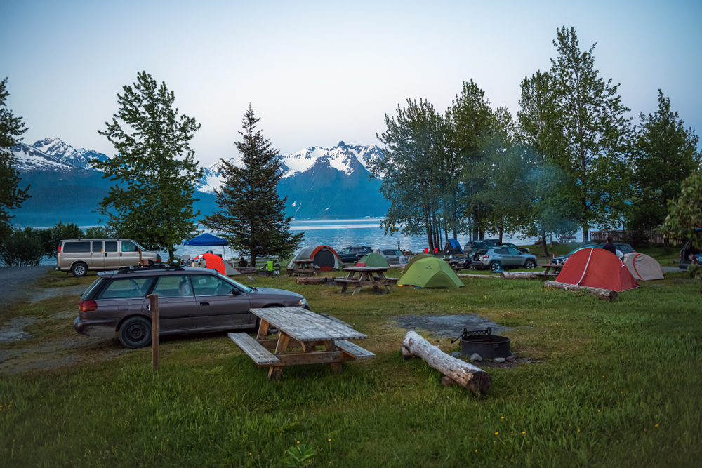 Camping in a park like setting along Resurrection Bay. There are tents and cars by the river with mountains in the background. 