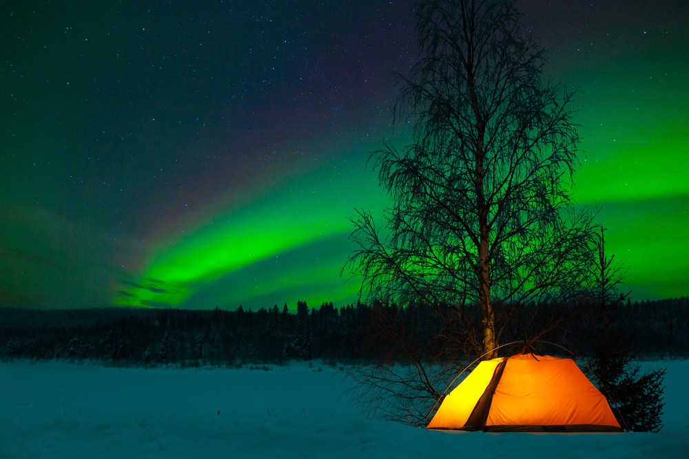 Camping in the north with the northern lights overhead. The tent is orange and the light about are green. The article is about camping in Alaska.   