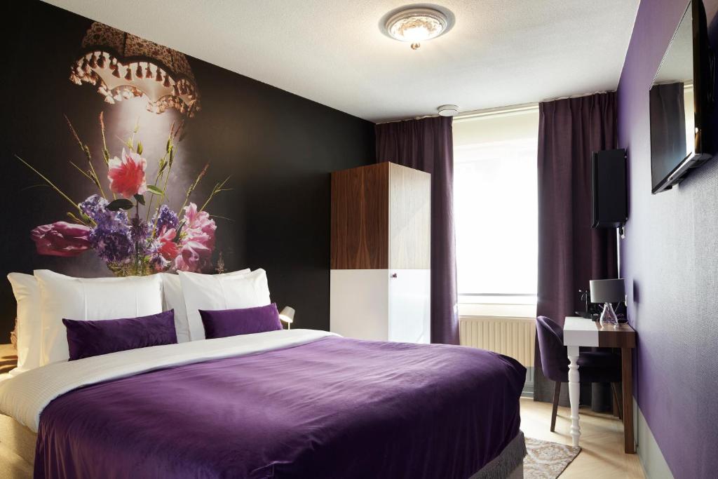 A small room with a purple and white bed, purple and black walls with a floral painting, and a large window