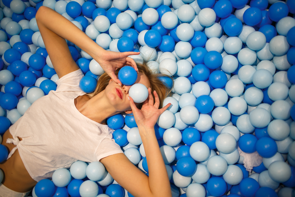 Young woman playing with balls in a dry pool. The balls are blue and white and she is holding two in front of here eyes. 