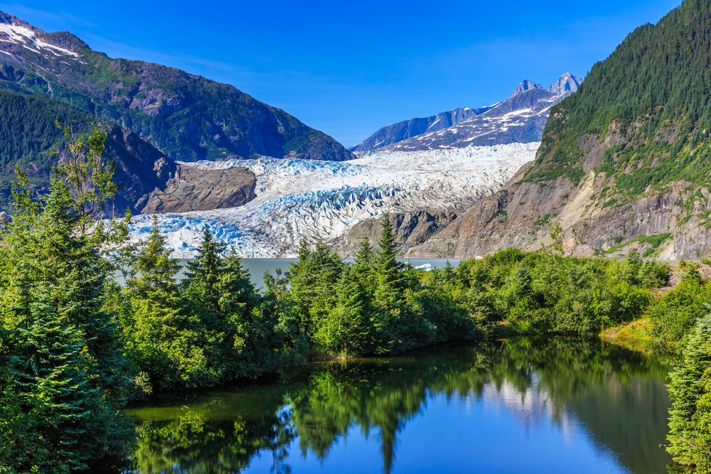 Sunny day at Mendenhall Glacier viewed across a lake and through evergreen trees.