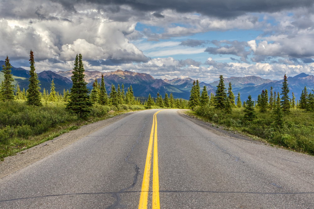 A paved highway going between trees toward the mountains.