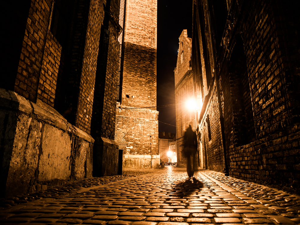 Illuminated cobbled street with light reflections on cobblestones in old historical city by night. Dark blurred silhouette of person evokes Jack the Ripper