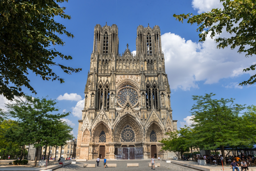 The front of the Gothic-style Reims Cathedral with tress around it.