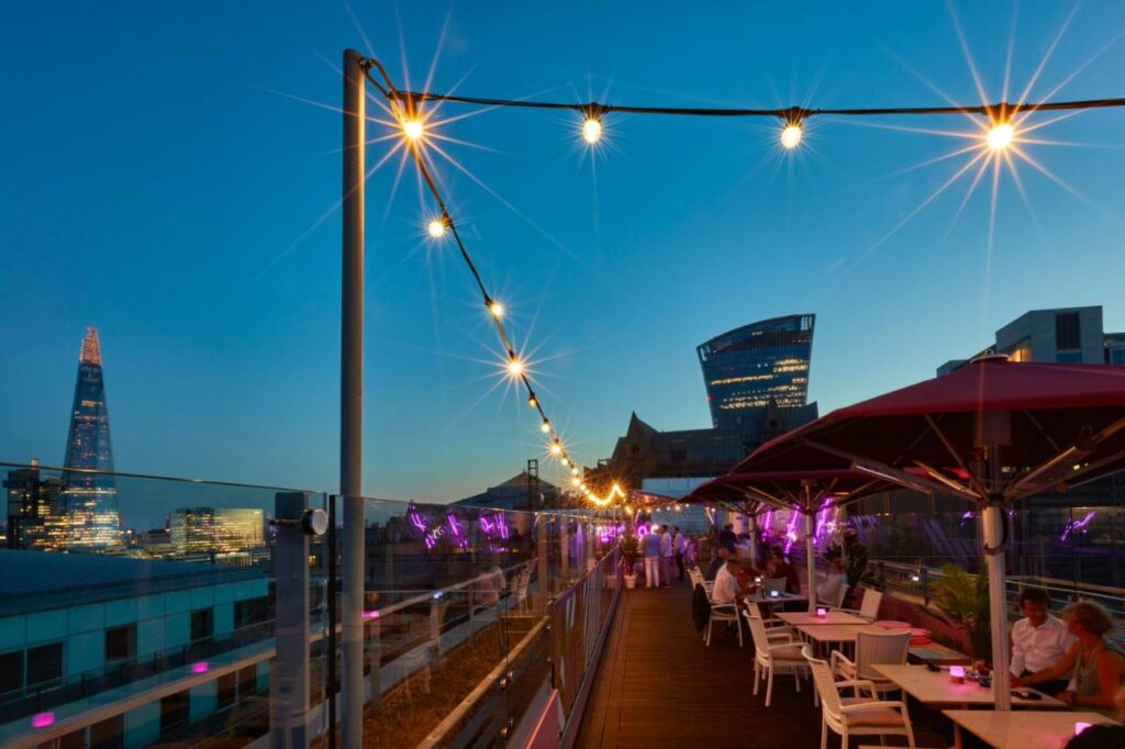Tables with umbrellas under a string of lights overlooking the city of London.