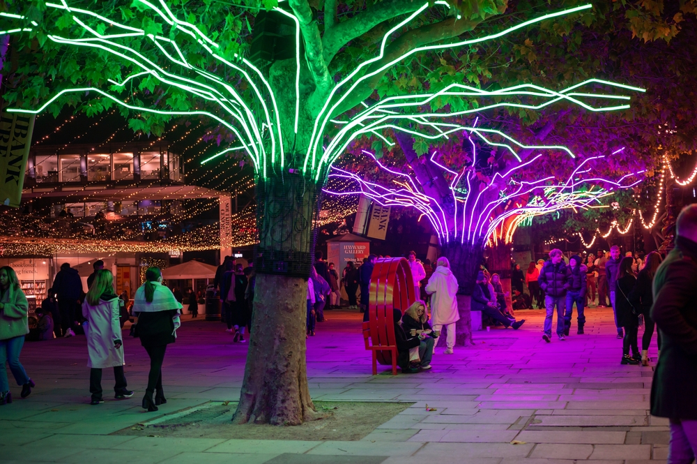 Shoppers enjoy the Southbank Christmas decorations at night. You can see market stalls in the background. 