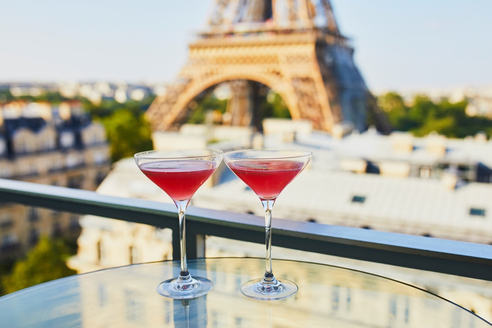 Two Cosmopolitan cocktails in traditional martini glasses on a glass table with view to the Eiffel tower, Paris, France. The article is about rooftop bars in Paris