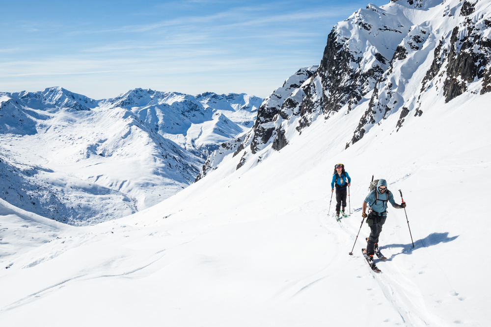 Two cross country skier on the side of a snowy mountain overlooking more mountains in Alaska in winter.