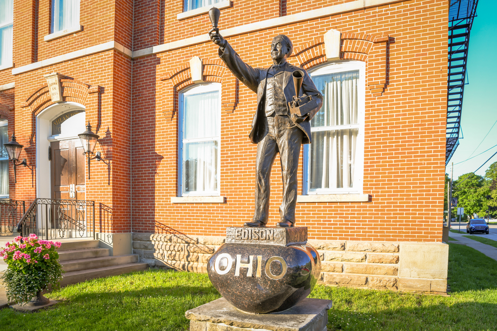 Statue of Thomas Edison holding a light bulb in front of a brick building.