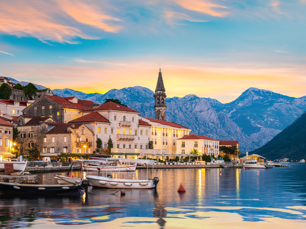 Pretty sunset over Perast and docked boats in the water with mountains in the distance.