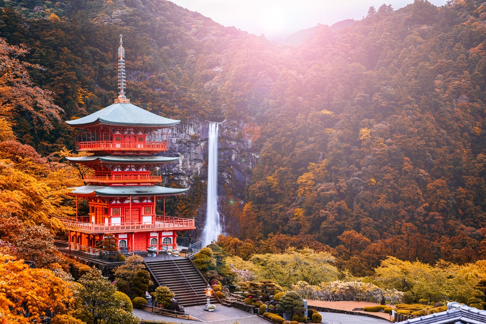 A tall red Japanese building in the valley of a mountain with a huge waterfall on the side of it and the trees are covered in fall leaves.