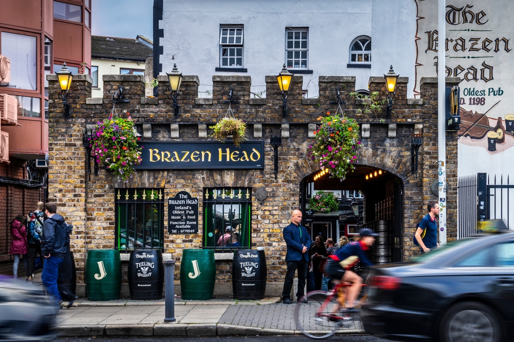 The Brazen Head, the oldest pub in Dublin. The brick pub is small with a turret like roof. there are people stood outdie the pub. 