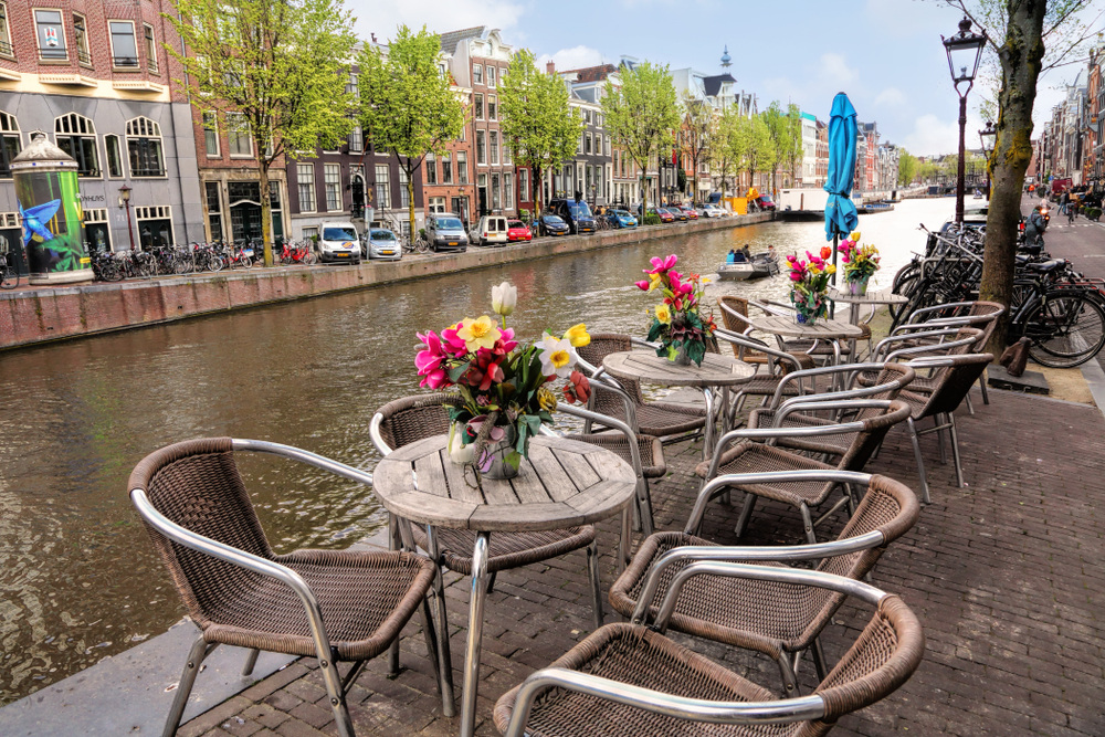 Small tables with chairs and flower vases lining a canal in Amsterdam.