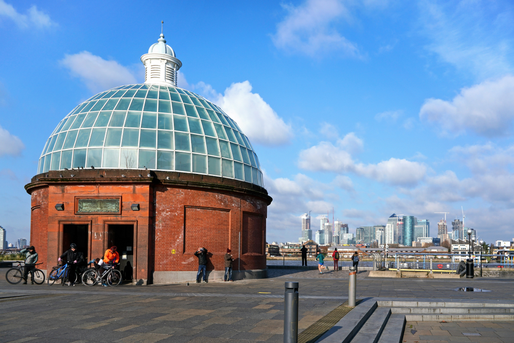 The entrance to the Greenwich Foot Tunnel, a round, brick building topped with glass, next to the river with the skyline in the distance.