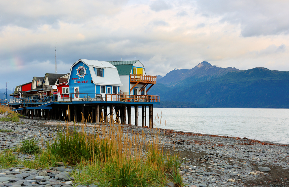 Colorful buildings on a raised boardwalk on the Homer Spit next to the water with mountains in the background.