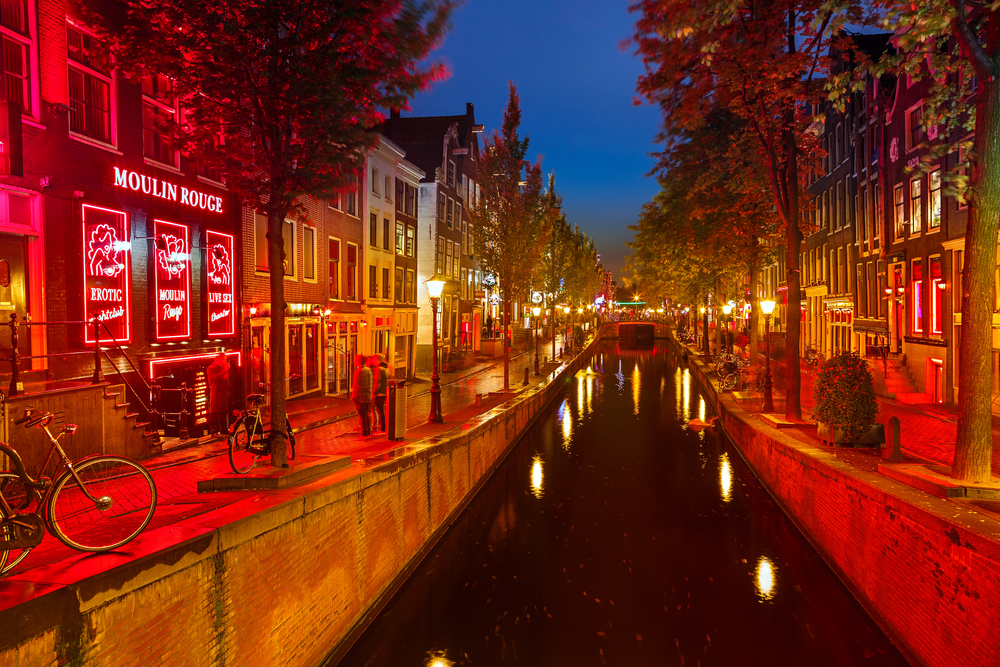 Canal cutting through the Red Light District at night with red-lit windows and the Moulin Rouge.