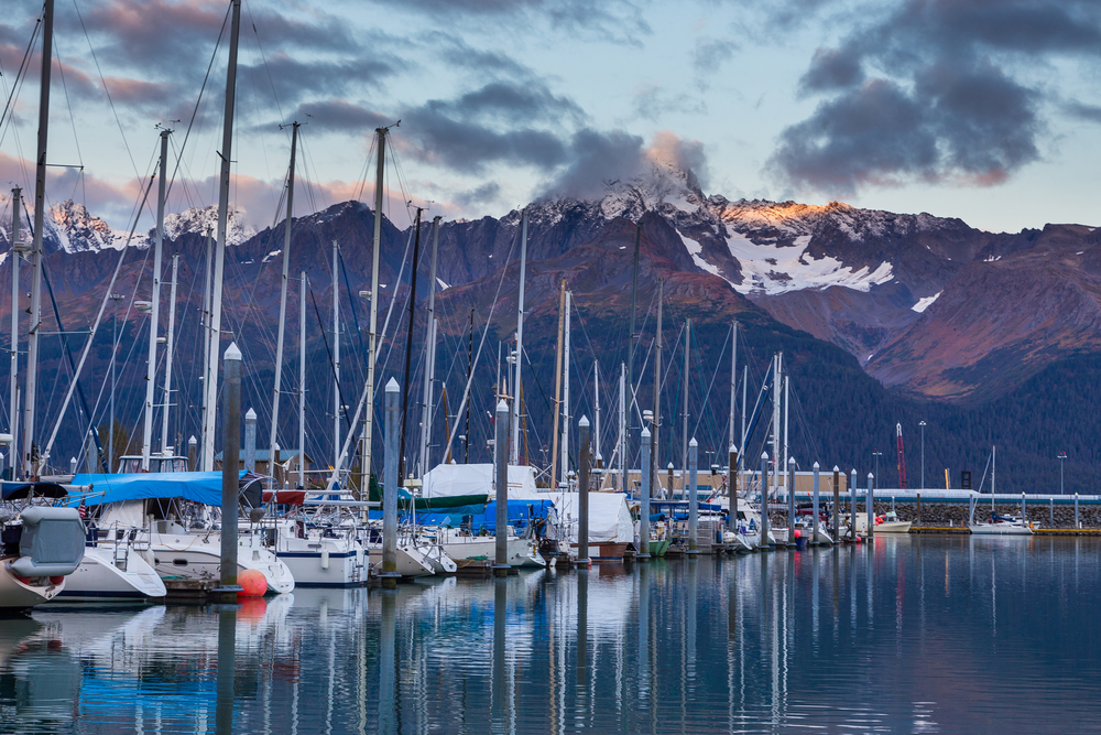 Line of boats docked in the Seward harbor at sunset with mountains in the distance.