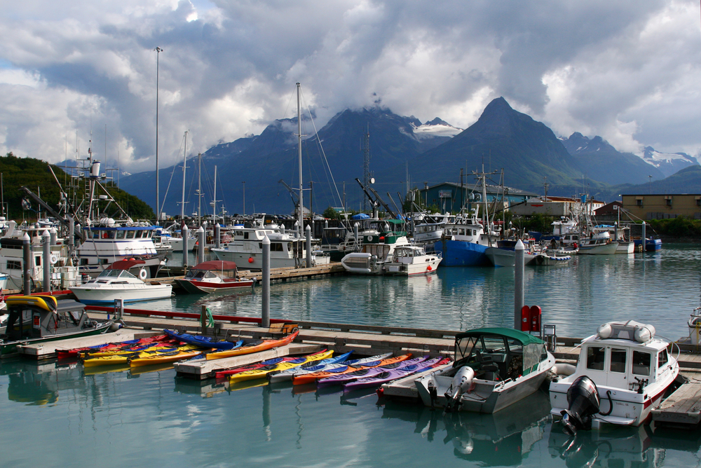 Harbor in Valdez full of docked boats and kayaks with cloud-covered mountains in the distance.