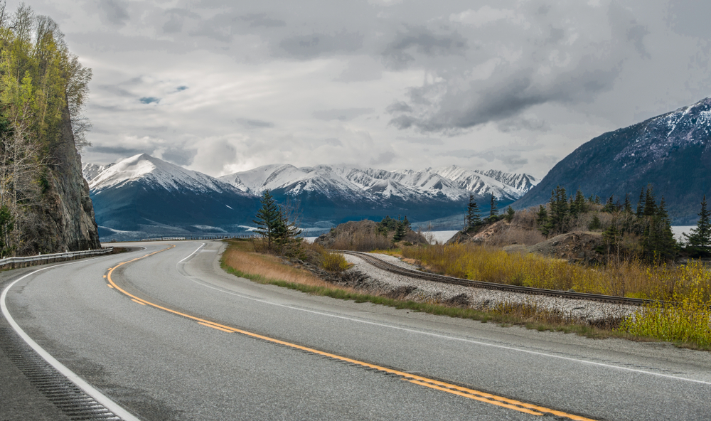 The Seward  Alaska Highway curves beneath cloudy skies as it passes by snow-covered mountains at the edge of an ocean inlet south of Anchorage.