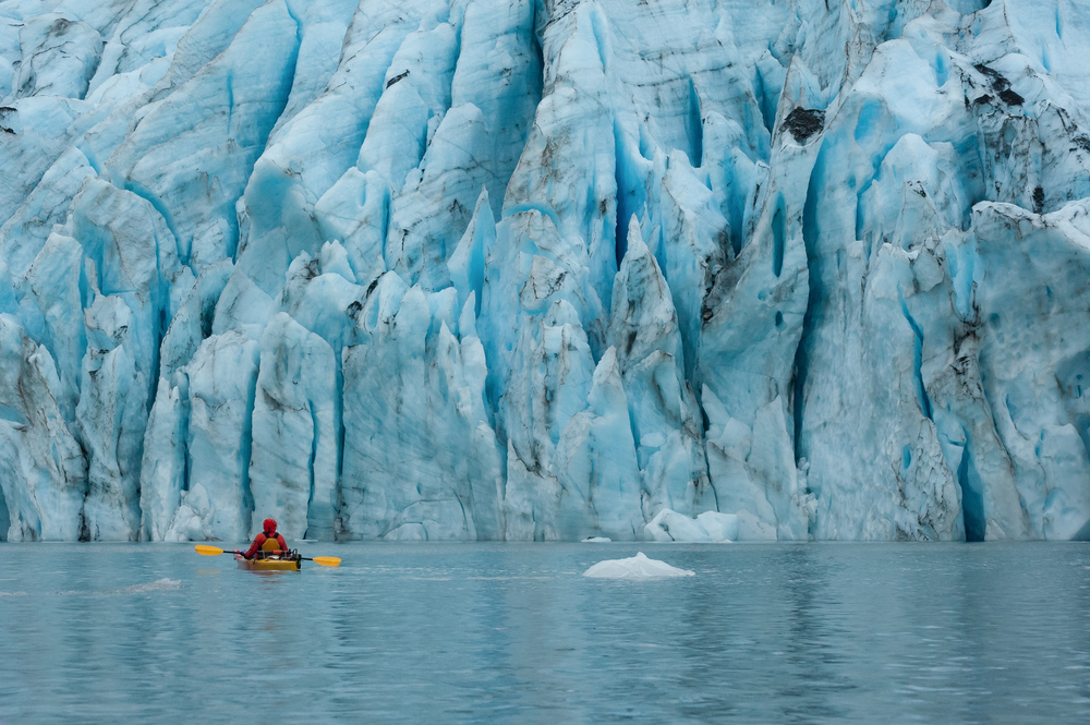 Single kayaker near the pale blue wall of rugged glacier.